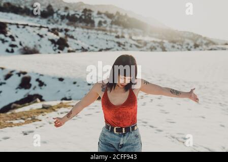 Giovane donna di stile rilassato con tatuaggi in top rosso e. jeans che si sparge le mani mentre si trova in una valle innevata in sole giorno invernale Foto Stock