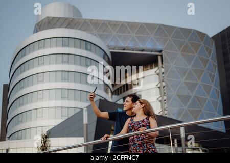 Romantica coppia in viaggio che prende selfie sul telefono cellulare in background di un incredibile edificio geometrico in balcone Foto Stock