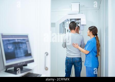 Vista posteriore del cliente che fa raggi X con l'aiuto di attento assistente in uniforme blu in cabina dentale con monitor e. attrezzatura Foto Stock