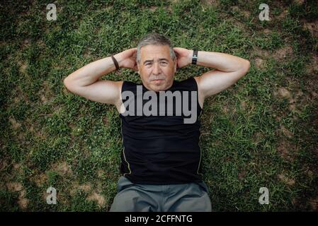 Da sotto forte uomo maturo attivo che guarda la macchina fotografica dentro buona forma sportiva facendo abs in prato verde nel parco in estate Foto Stock