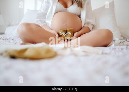 Crop femmina incinta senza volto vestita in blusa bianca aperta e. bra mostrando abiti per non nato mentre si siede a letto con gambe incrociate Foto Stock