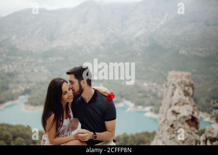Donna felice che trasporta il fiore mentre l'uomo bacia la guancia e si siede alla cima della montagna rocciosa con vista panoramica Foto Stock
