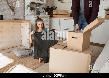 Felice giovane coppia che prende fuori le maglie e le cose grigie morbide in scatole di cartone in cucina bianca Foto Stock