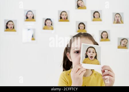 Carino bambina in abito giallo nascondendo faccia dietro di sé immagini mentre si è in piedi contro il muro con foto che dimostrano varie emozioni Foto Stock