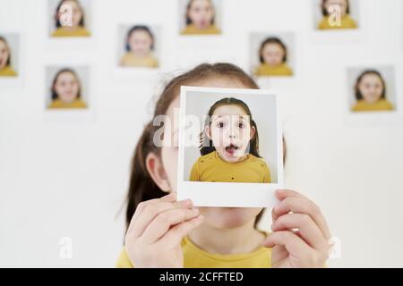 Carino bambina in abito giallo nascondendo faccia dietro di sé immagini mentre si è in piedi contro il muro con foto che dimostrano varie emozioni Foto Stock