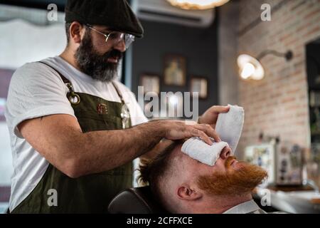 Barbiere facendo massaggio viso a bel rosso uomo con chiuso occhi seduti in sedia con un caldo asciugamano che copre la faccia Foto Stock