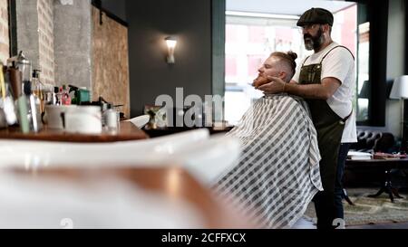 Barbiere facendo massaggio viso a bell'uomo rosso con seduta in sedia Foto Stock