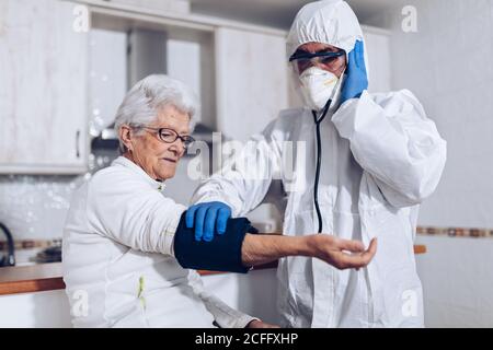 Assistente professionale di cura della casa in uniforme protettiva e maschera che esamina donna anziana e controllo della pressione sanguigna durante la visita del paziente a casa durante l'epidemia di coronavirus Foto Stock