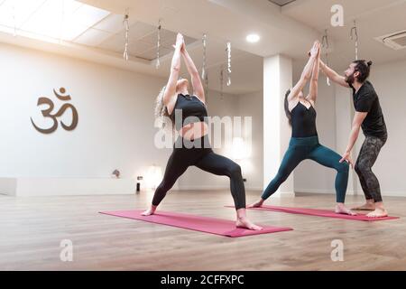 Equilibrato calma donne e insegnante di uomo che aiutano a correggere pose mentre si allungano e alzano le mani in su in una postura guerriero in corso di yoga Foto Stock