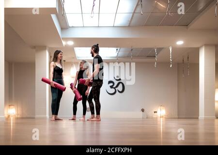 Giovane uomo e donna con tappetini che parlano l'uno con l'altro mentre si è in piedi in uno spazioso studio durante l'allenamento di yoga Foto Stock