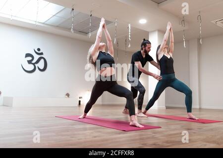 Equilibrato calma donne e insegnante di uomo che aiutano a correggere pose mentre si allungano e alzano le mani in su in una postura guerriero in corso di yoga Foto Stock