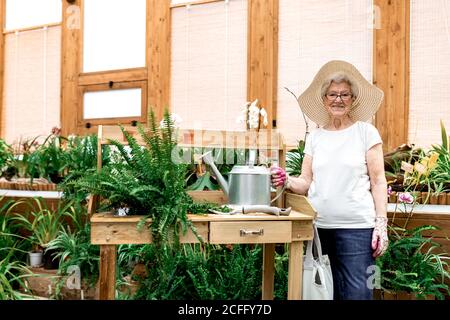 Gentile signora anziana con annaffiatura può sorridere e guardare fotocamera in piedi vicino al tavolo in un attico di legno Foto Stock
