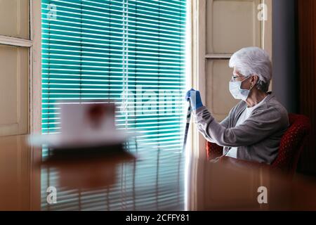 Donna anziana in maschera protettiva e guanti con bastone da passeggio seduto vicino alla finestra Foto Stock