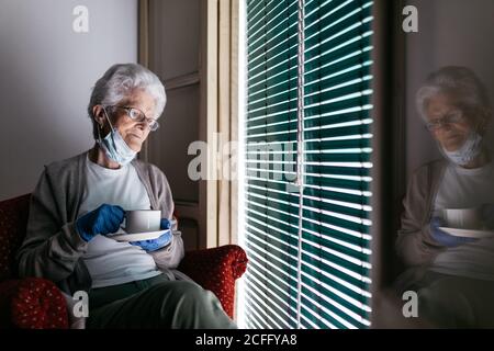 Triste pensiva vecchia femmina con maschera protettiva e guanti seduta vicino alla finestra con una tazza di tè in mano trascorrere una giornata da sola a casa durante la pandemia del coronavirus Foto Stock