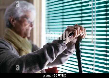 Vista laterale di una donna anziana pensiva con una canna da passeggio seduto vicino alla finestra con le porte e guardando lontano mentre si passa la giornata da solo a casa Foto Stock