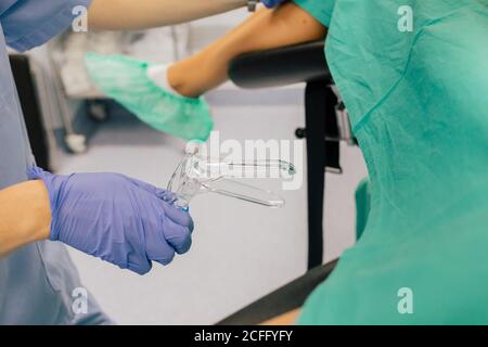 Medico femminile senza volto in guanti blu e uniforme con plastica specchio ginecologico che esamina pazienti anonimi in copriscarpe su cui giace sedia in clinica di fertilità Foto Stock