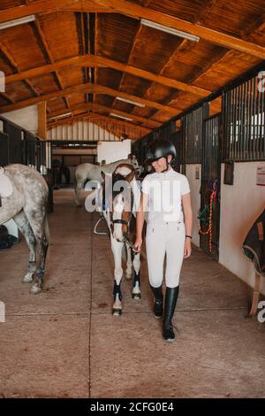 Giovane donna adolescente in vestito bianco e casco jockey leader cavalcare fuori dalla stalla per andare a cavallo all'esterno