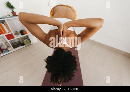 Vista posteriore dell'anonima giovane donna afroamericana attraente che esegue la postura di yoga con le mani ripiegate dietro la schiena in stanza leggera Foto Stock