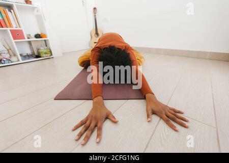 Giovane donna anonima afroamericana che esegue posa yoga con testa giù e mani gambe che si allungano sul tappetino in stanza leggera Foto Stock