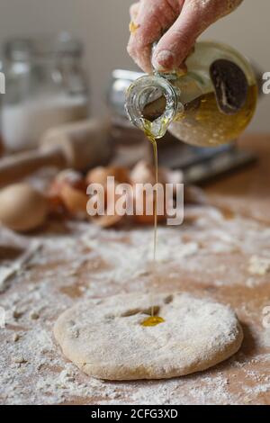 cuocete senza volto versando olio d'oliva fresco e fragrante dalla caraffa di vetro su pasta arrotolata su sfondo di pasticcio di cucina sfocato Foto Stock