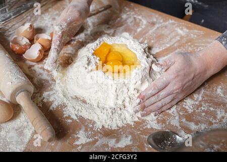 persona anonima in vestiti neri che fanno recesso in massa di farina con la mano per aggiungere le uova mentre si cucinano i dolci per la pasta a casa Foto Stock