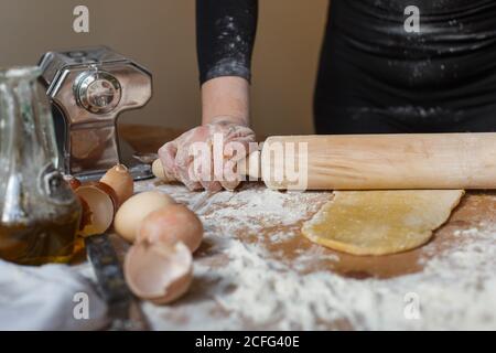 Donna anonima in abito nero arrotolando l'impasto con legno tondino sul tavolo con farina e vari utensili da cucina mentre si prepara la pasta Foto Stock