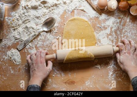 Donna anonima in abito nero arrotolando l'impasto con legno tondino sul tavolo con farina e vari utensili da cucina mentre si prepara la pasta Foto Stock