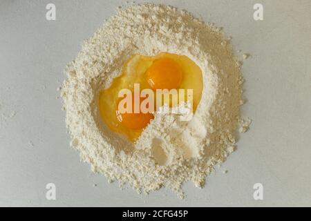 Vista dall'alto del cumulo di farina con uova grezze rotte tavolo da cucina centrale in legno con ingredienti di pasta e cucina utensil in giorno di sole luminoso Foto Stock