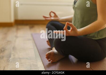 Vista laterale di una donna anonima in abbigliamento sportivo seduto a piedi nudi Pavimento e facendo yoga esercizio in Padmasana con mudra gesto Foto Stock