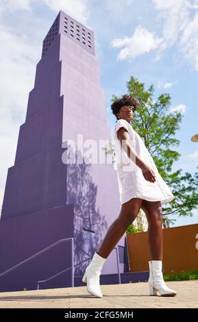 Dal basso vista di Snella African American Woman in abito bianco e stivali che si posano da soli contro l'edificio a più piani viola in città in sole giornate estive Foto Stock