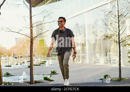 Allegro uomo ispanico con cuscinetti in abiti casual sorridente e guardando con occhiali da sole e cuffie alla moda sul collo in città via Foto Stock