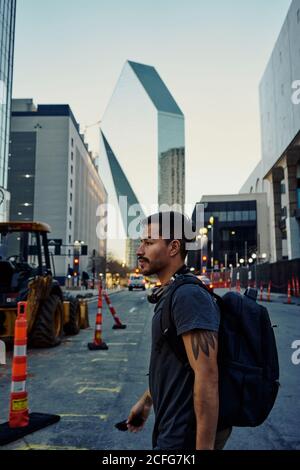 Vista laterale del viaggiatore ispanico maschio con abbigliamento casual zaino e cuffie sul collo camminando lungo la moderna strada della città Foto Stock