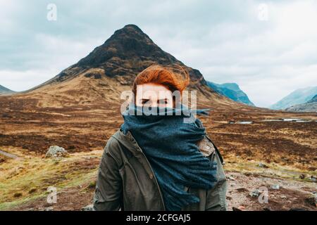 Bella zenzero Donna swaddled in sciarpa in piedi contro le montagne pittoresche della Scozia Foto Stock