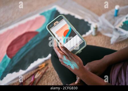 Ritagliare una donna afroamericana irriconoscibile utilizzando un'app con un campione di immagine sullo smartphone mentre si impara a dipingere a casa durante il tempo libero Foto Stock
