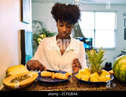 Donna afroamericana in camicia casual seduta a tavola con vari frutti tropicali e papaya tritare Foto Stock