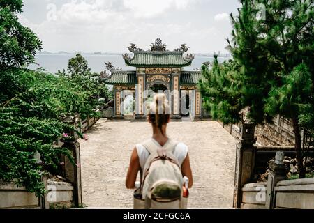 Vista posteriore di turista femminile anonimo con zaino in piedi scale di fronte all'arco del tempio buddista in Vietnam Foto Stock