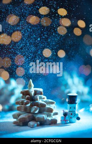 Simpatico pupazzo di neve e abete dal biscotto sulla neve meteo con luci su sfondo sfocato Foto Stock