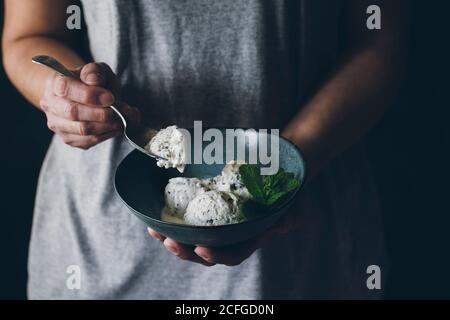 Vista in pianta della donna anonima con cucchiaio in mano e ciotola con palline di gelato alla stracciatella decorate con foglie di menta Foto Stock