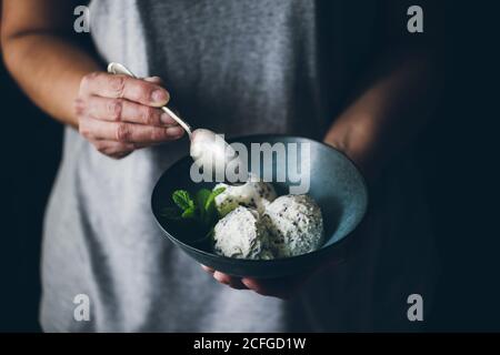 Vista in pianta della donna anonima con cucchiaio in mano e ciotola con palline di gelato alla stracciatella decorate con foglie di menta Foto Stock