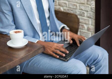 foto senza volto di uomo nero in tuta blu seduto con una tazza di caffè al tavolo e il computer portatile di navigazione in ginocchio. Foto Stock