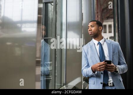 Uomo nero serio in tuta trendy che tiene smartphone e guardando via con attenzione attraverso il vetro. Foto Stock
