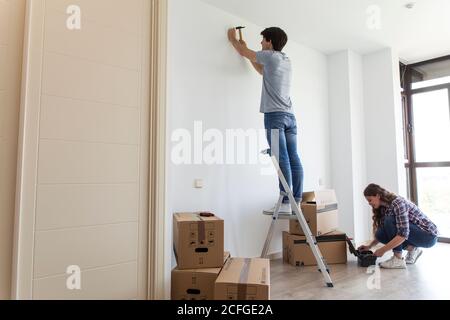 Giovane uomo in piedi sulla scala martellando chiodo e Donna raccogliendo strumenti dalla borsa in appartamento con scatole di cartone Foto Stock