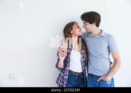 Giovane coppia in abbigliamento casual baciare e tenere le mani in piedi in camera con parete bianca vuota Foto Stock