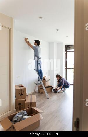 Giovane uomo in piedi sulla scala martellando chiodo e Donna raccogliendo strumenti dalla borsa in appartamento con scatole di cartone Foto Stock