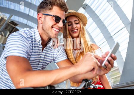 Bel giovane e bella Donna sorridente e navigante smartphone moderno, mentre si appoggia in bicicletta nel sentiero coperto nelle giornate di sole Foto Stock