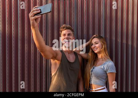 Attraente giovane uomo e donna sorridente e in posa per selfie mentre si trova vicino al muro sulla strada Foto Stock