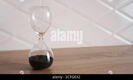 Clessidra di cristallo con sabbia nera su tavolo di legno e bianco sfondo Foto Stock