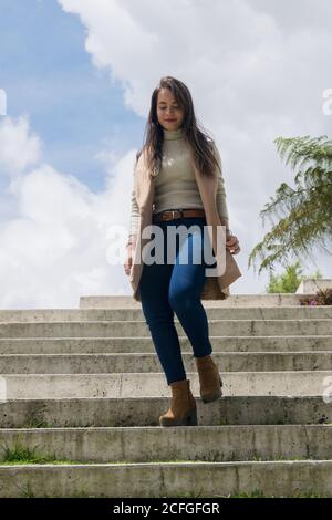 Da sotto giovane attraente capelli lunghi Donna in dolcevita chiaro e giacca senza maniche che cammina giù scale di pietra in giorno nuvoloso Foto Stock