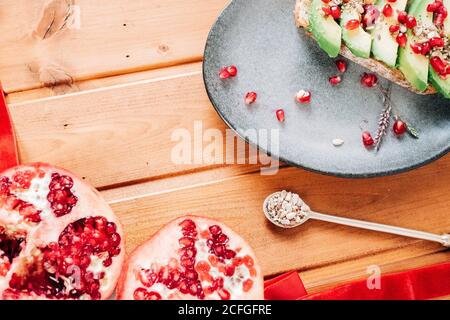 Vista dall'alto del melograno dimezzato e del cucchiaio con i semi inseriti vicino piatto con delizioso toast avocado su tavolo di legno Foto Stock