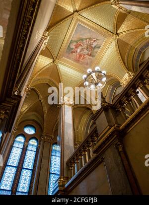 Opulento soffitto parlamentare: Una candelabra dorata illumina il soffitto dipinto incorniciato da foglie d'oro in una scala dell'edificio del Parlamento ungherese. Foto Stock